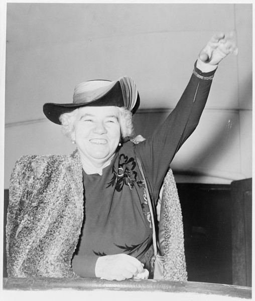 Fotografia em preto de branco de Elizabeth Kenny sorrindo e levantando o braço esquerdo. Fotografia em preto de branco de Elizabeth Kenny sorrindo e levantando o braço esquerdo.